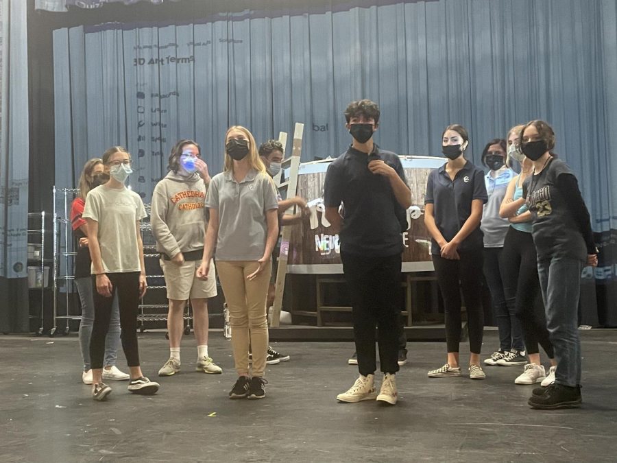 Drama production cast working on choreography with tech crew working on the set behind them. Ms. Wilson is giving them pointers to remember their placements. 