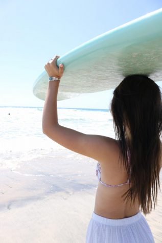Mia Boggs carrying her surfboard on top of her head while looking out into the distance. She gazes at the blue ocean water that glitters because of the bright and blazing rays of the sun.