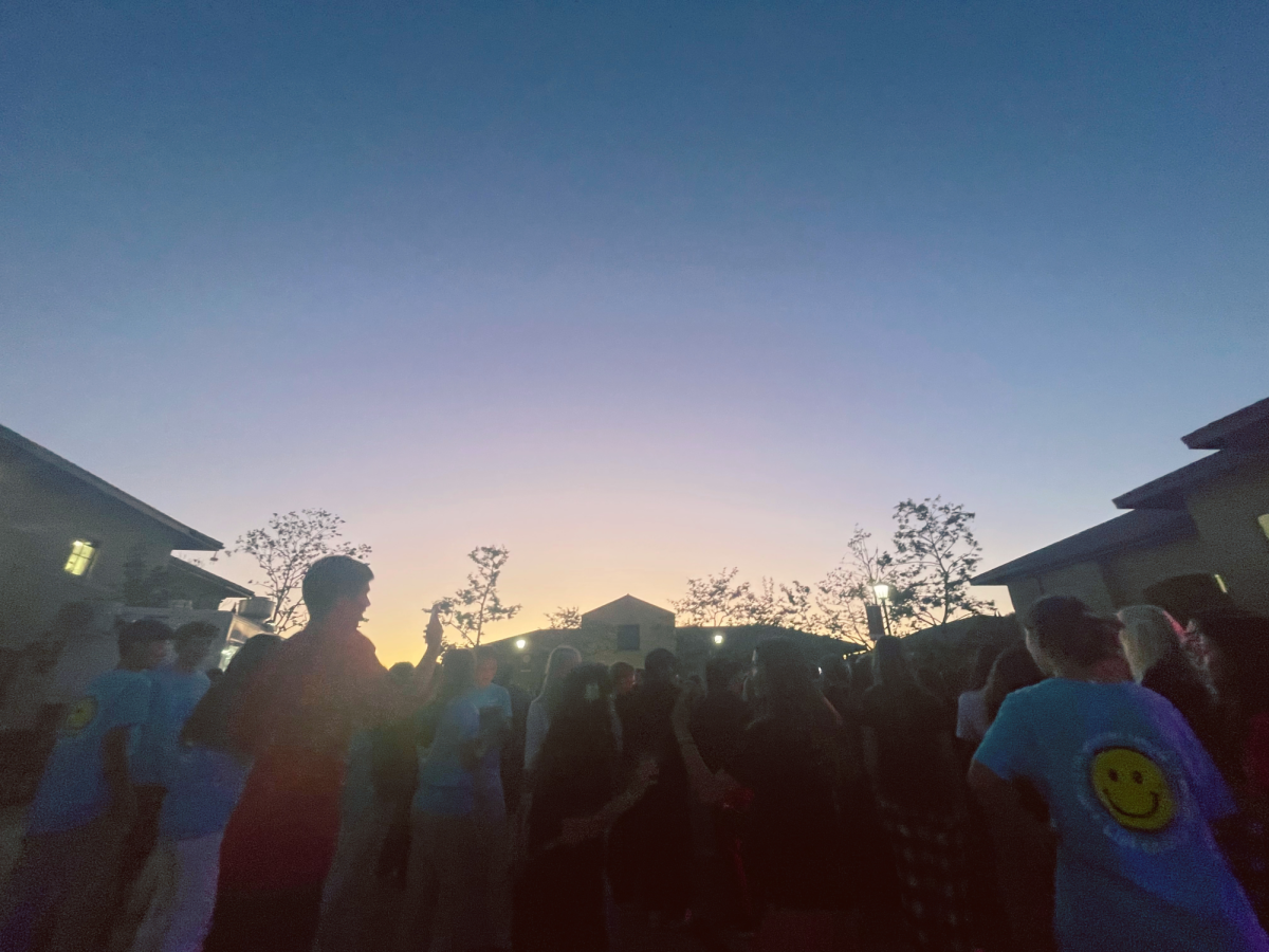 A large crowd of kids at a school dance; they’re standing in a school courtyard at sunset, and many are holding up phones or jumping up and down